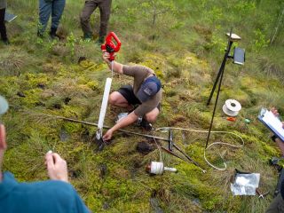 Pētnieks purvā uzstāda gruntsūdens monitoringa cauruli un veic mērījumus ar elektronisku ūdens līmeņa mērītāju. Blakus novietotas monitoringa iekārtas, sensori un instrumenti, bet apkārt esošie dalībnieki novēro darbu un fiksē rezultātus