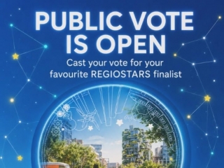 Alt teksts: Plakāts ar aicinājumu balsot REGIOSTARS 2026 konkursā. Uz zila fona redzams teksts “PUBLIC VOTE IS OPEN – Cast your vote for your favourite REGIOSTARS finalist”. Centrā apaļā ilustrācijā attēlota mūsdienīga, zaļa pilsētvide ar gājējiem, sabie